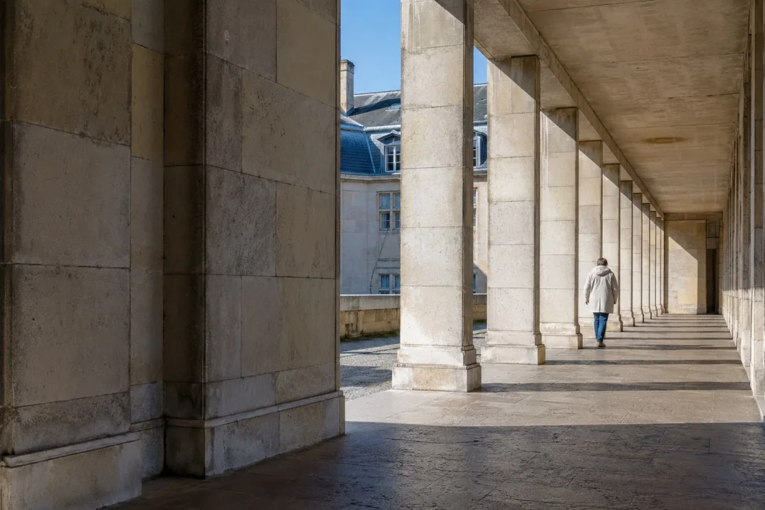 A limestone colonnade with a figure in a light coat walking away beneath the arcade, a mansard-roofed building visible beyond the columns.